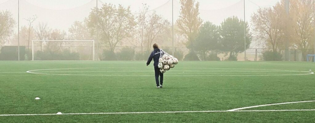 A person with a net full of soccer balls walking down a soccar field with the goal in the distance